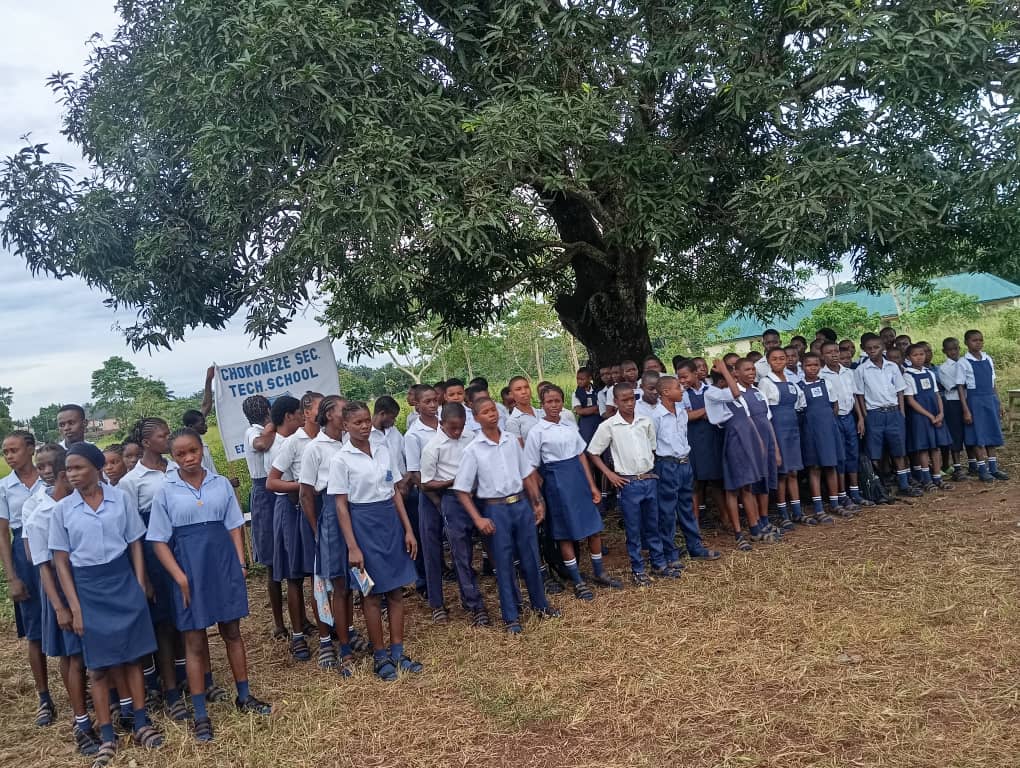 Chokoneze Secondary Technical School students assembled under the shade of the mango tree with school banner in blue skirts/trousers and white shirts